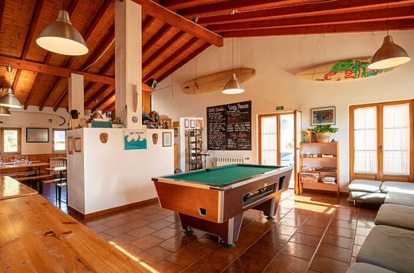 alt="Salón social del Russo Surfcamp con mesa de billar, tablas de surf decorativas y zona de bar acogedora con techos de madera"