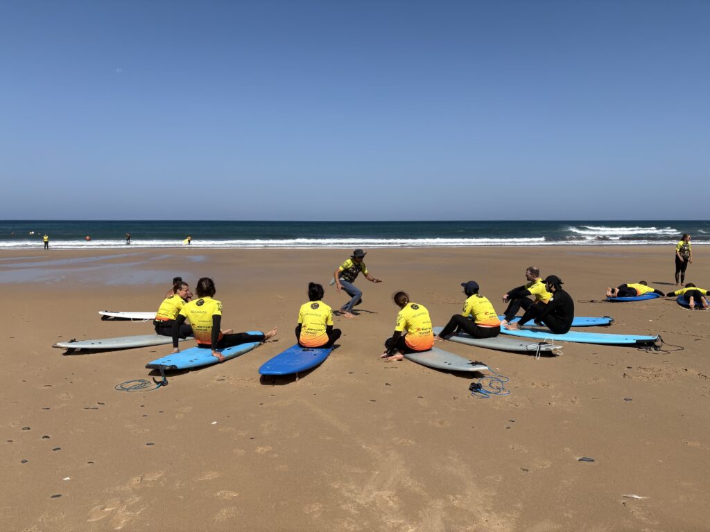 alt="Monitor de surf dando explicaciones técnicas a un grupo de alumnos sentados en la arena"