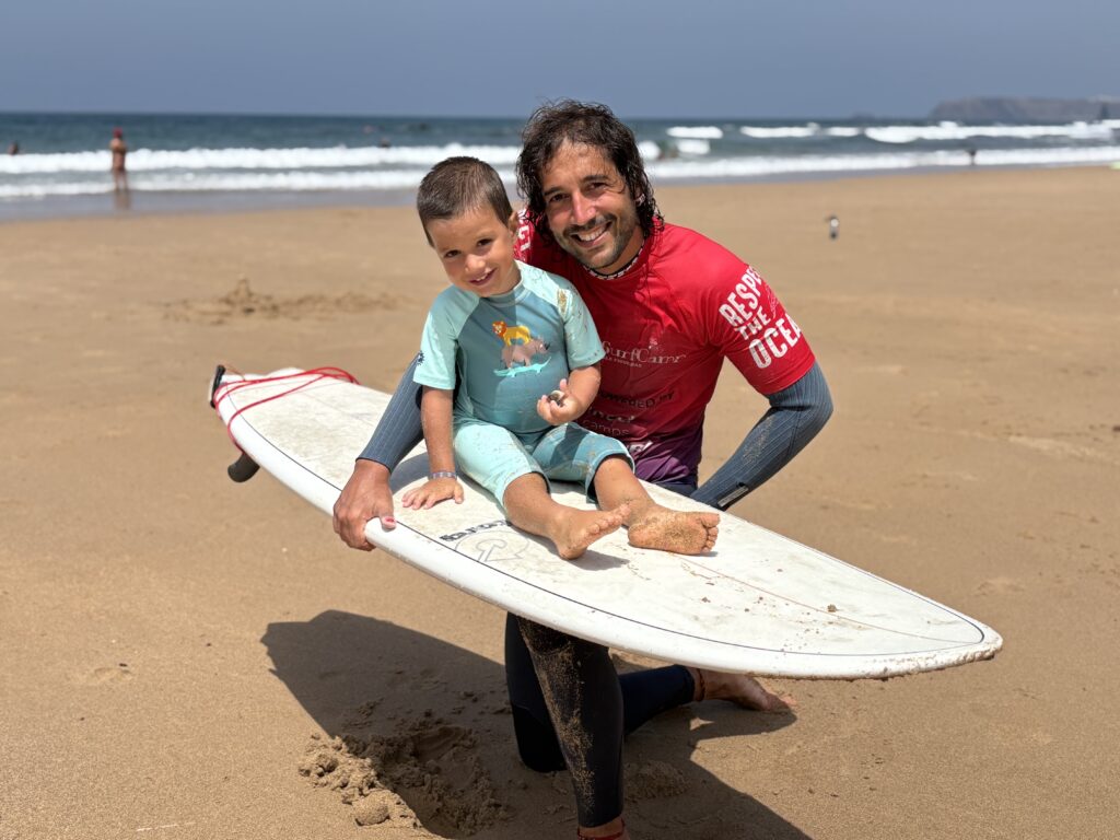 alt="Monitor de surf sujetando a un niño pequeño sentado sobre una tabla en la orilla del mar"