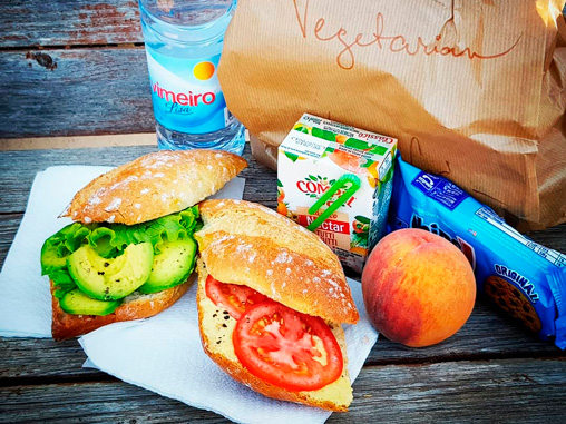 alt="Pack de picnic para la playa con sándwiches de aguacate y tomate, fruta, zumo y agua sobre madera"
