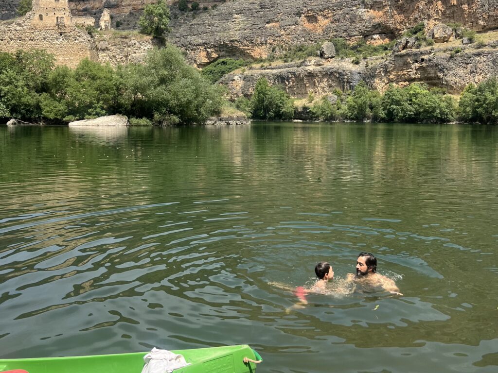 Padre e hijo dándose un baño en las aguas verdes del río Duratón con los cañones y ruinas de fondo durante la excursión por las Hoces del Duratón con niños.